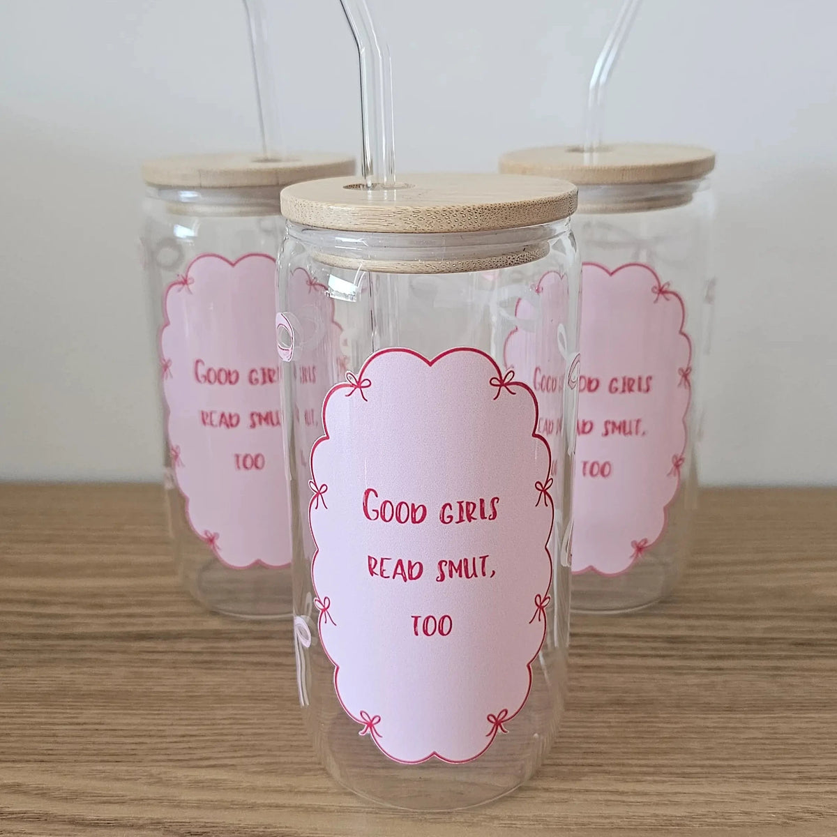 Three 16oz 'Good Girls Read Smut, Too' glass can tumblers with bamboo lids and glass straws, featuring a pink scalloped label with bow details, displayed on a wooden surface