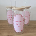 Three clear glass mugs with bamboo lids and clear bent straws, arranged on a wooden surface against a plain light wall. The front mug has a pink scalloped label that reads 'Good girls read smut, too'.
