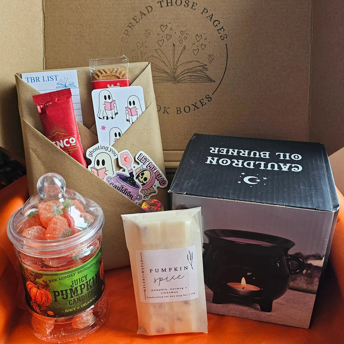 A curated gift box or subscription box from 'SPread Those Pages Book Boxes' containing various items: a jar of orange gummy candies, a pumpkin spice wax melt, a blind date with a book, a black witches cauldron wax burner, all arranged inside a brown cardboard box with an orange interior.