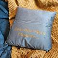 A square blue throw pillow with gold embroidered text reading 'Just One More Chapter.