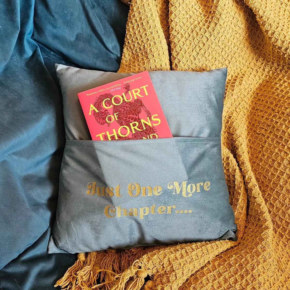 A slate blue decorative pillow with gold embroidered text reading 'Just One More Chapter, holding an ACOTAR book in it's front pocket 