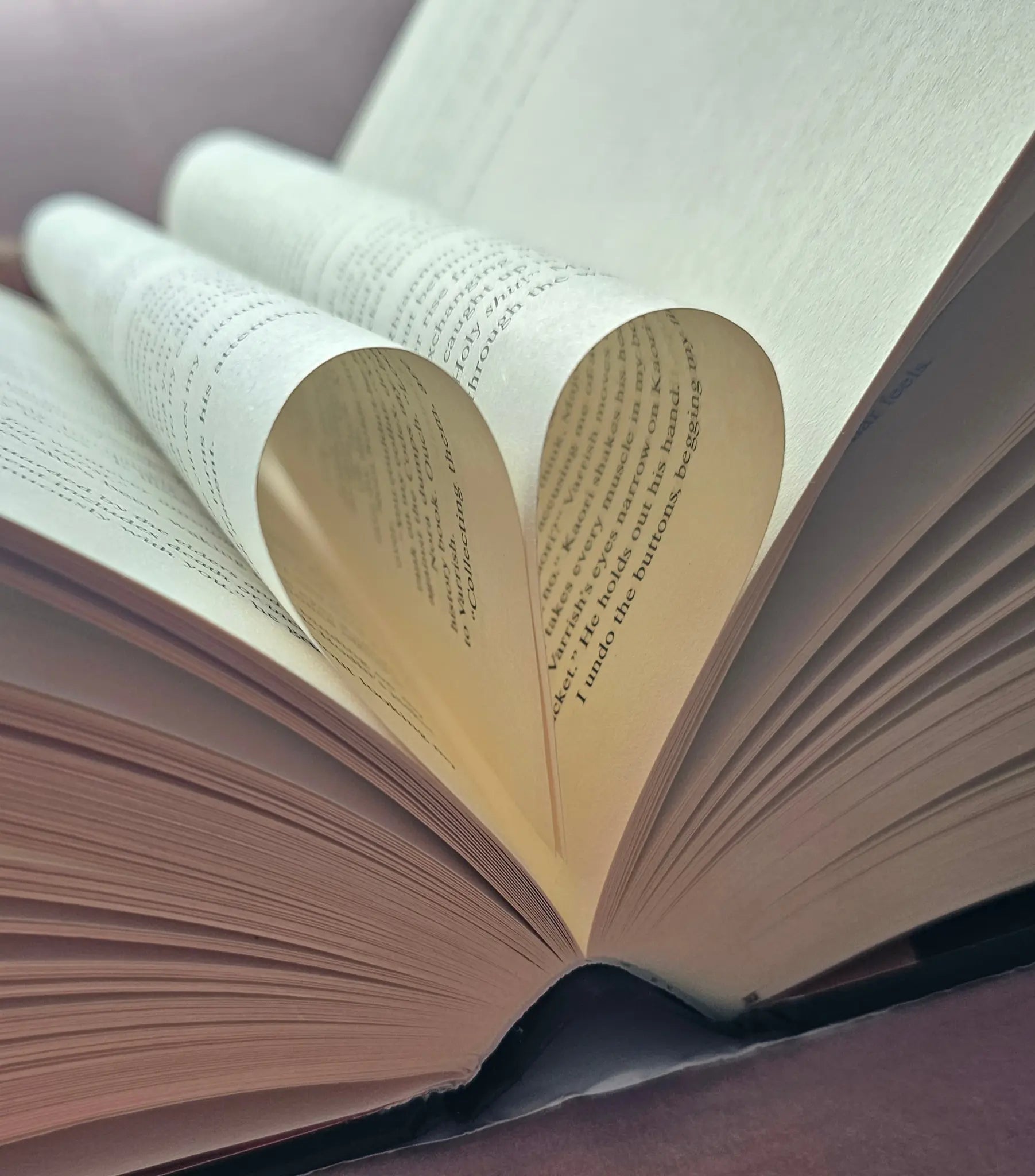 An open fantasy romance hardback book with pages curled into a heart shape, photographed close up against a soft purple background.