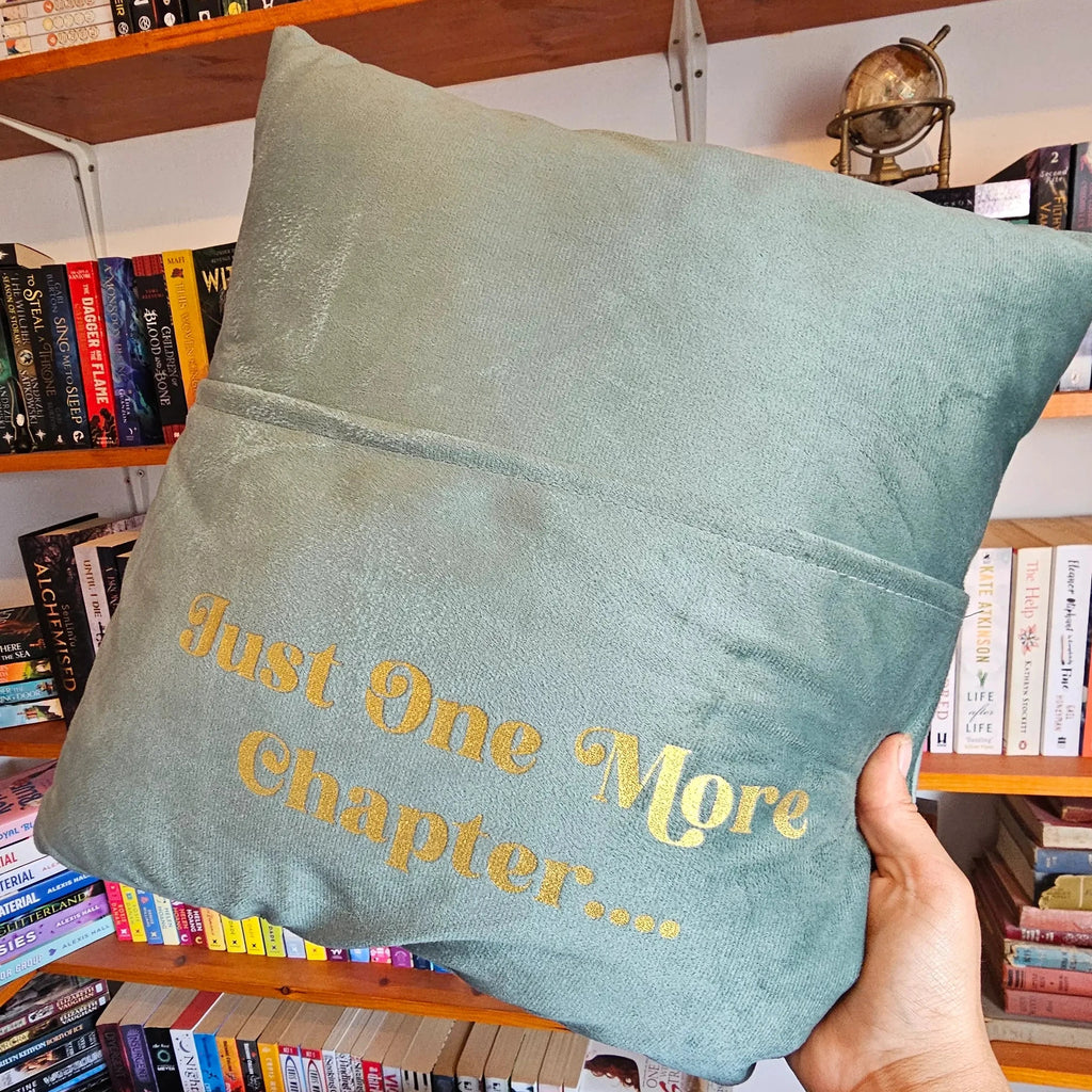 Slate blue velvet cushion with gold Just One More Chapter lettering and book pocket, held up in front of a bookshelf