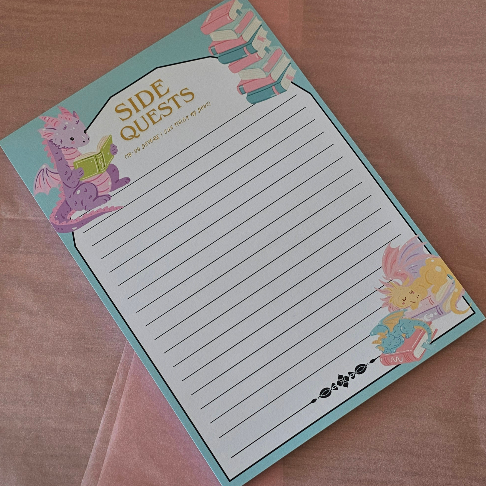 A 'Side Quests' lined notepad with a light blue border and whimsical fantasy illustrations of a purple dragon and a yellow rabbit reading books, displayed on a pink fabric background.