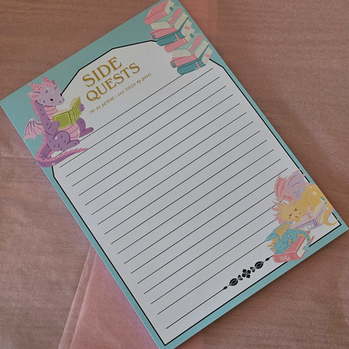 A 'Side Quests' lined notepad with a light blue border and whimsical fantasy illustrations of a purple dragon and a yellow rabbit reading books, displayed on a pink fabric background.