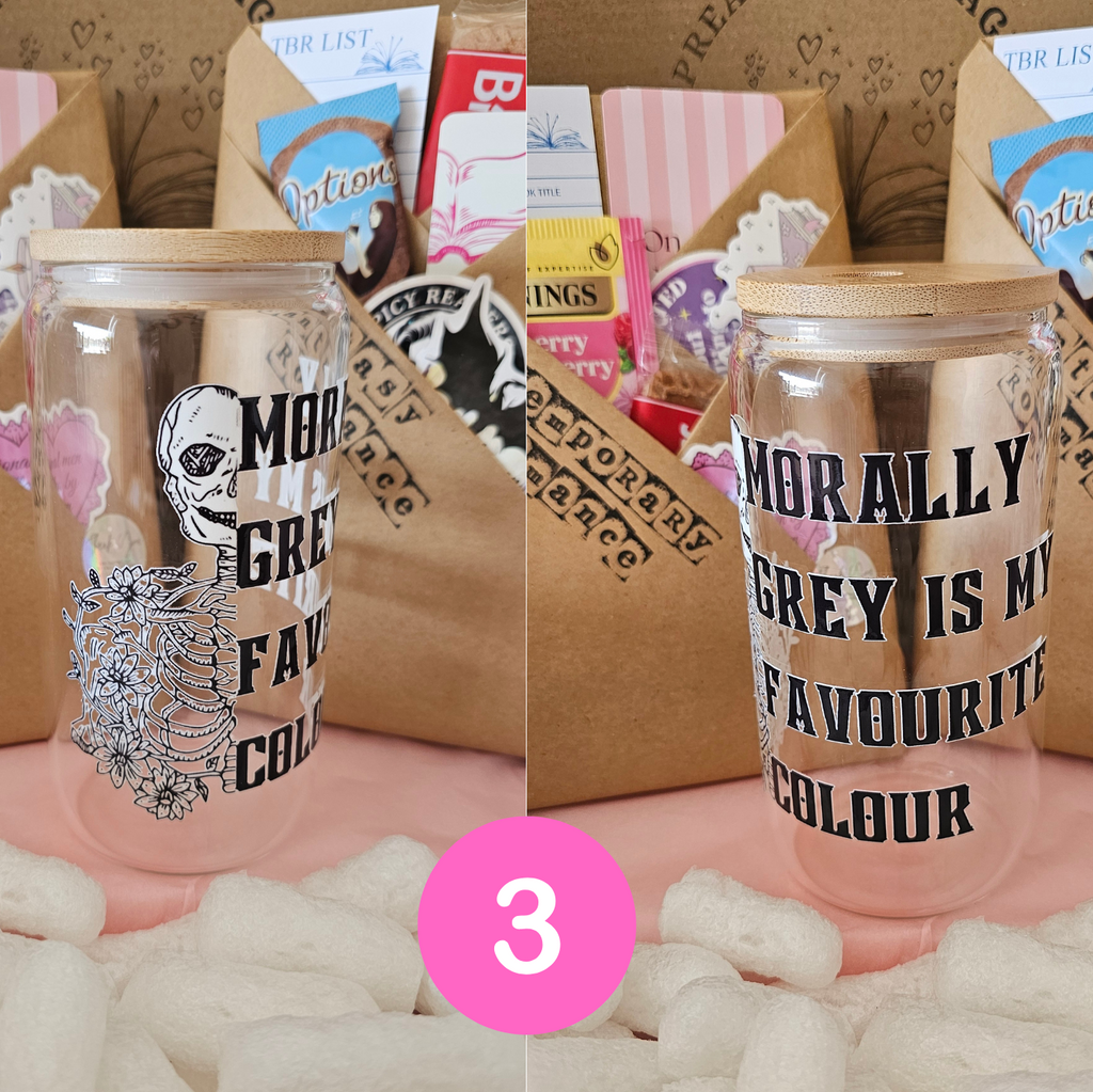 A clear glass can-shaped tumbler with a light wood lid, printed with the phrase 'Morally grey is my favourite colour' and a skull illustration, shown in front of a gift box with snacks. A pink circle with the number 3 is overlaid at the bottom.