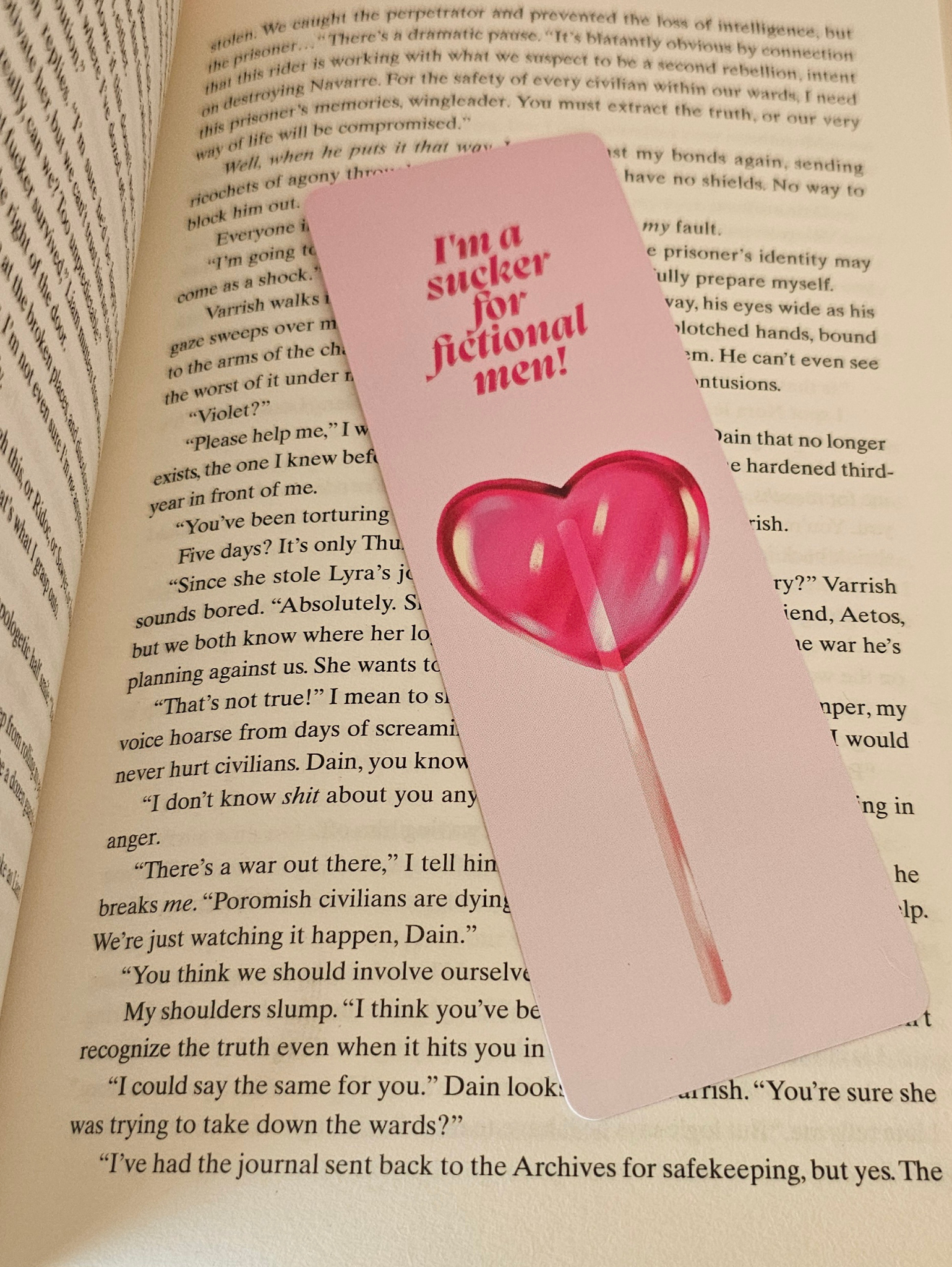 A pink paper bookmark with a red heart and the text 'I'm a sucker for fictional men!' resting on open book pages.