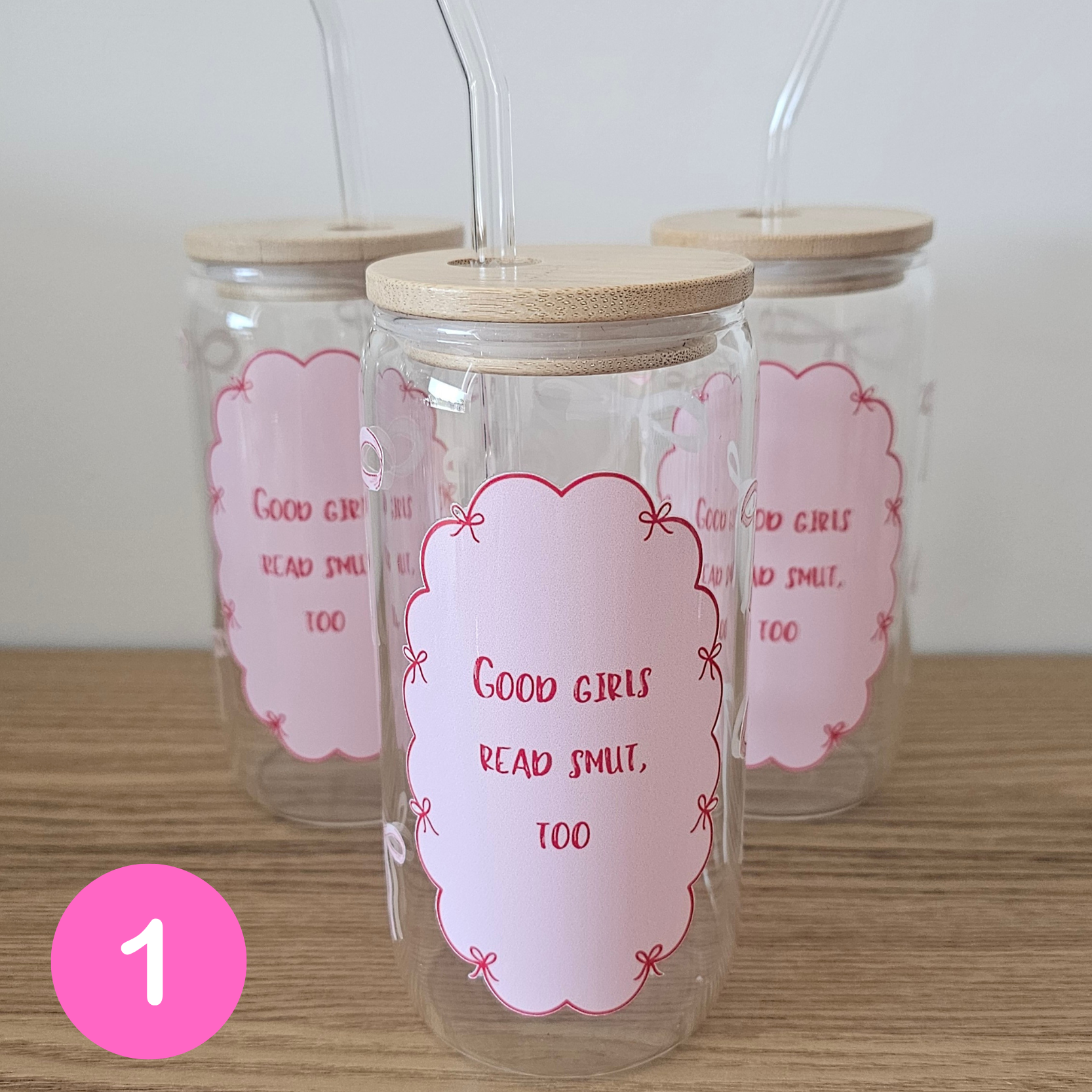 Three clear glass tumblers with bamboo lids and clear bent glass straws, decorated with pink labels that read 'Good girls read smut, too'. A pink circle with the number 1 is overlaid in the bottom-left corner.