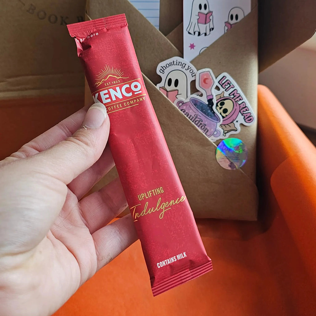 "Hand holding a Kenco Uplifting Indulgence Pumpkin spiced lattee coffee sachet from a Spread Those Pages Halloween book box, with ghost stickers and kraft paper packaging visible in the background"