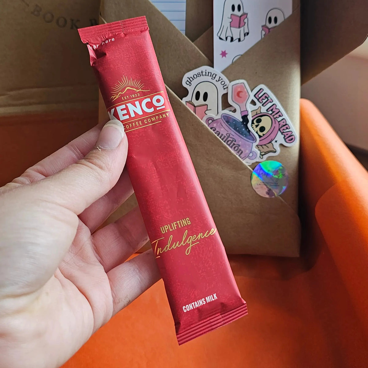 "Hand holding a Kenco Uplifting Indulgence Pumpkin spiced lattee coffee sachet from a Spread Those Pages Halloween book box, with ghost stickers and kraft paper packaging visible in the background"