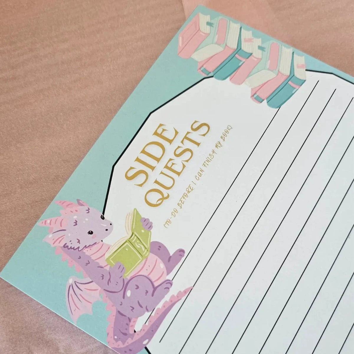 "Side Quests dragon notepad angled view showing purple reading dragon illustration, gold title and lined pages on teal cover"