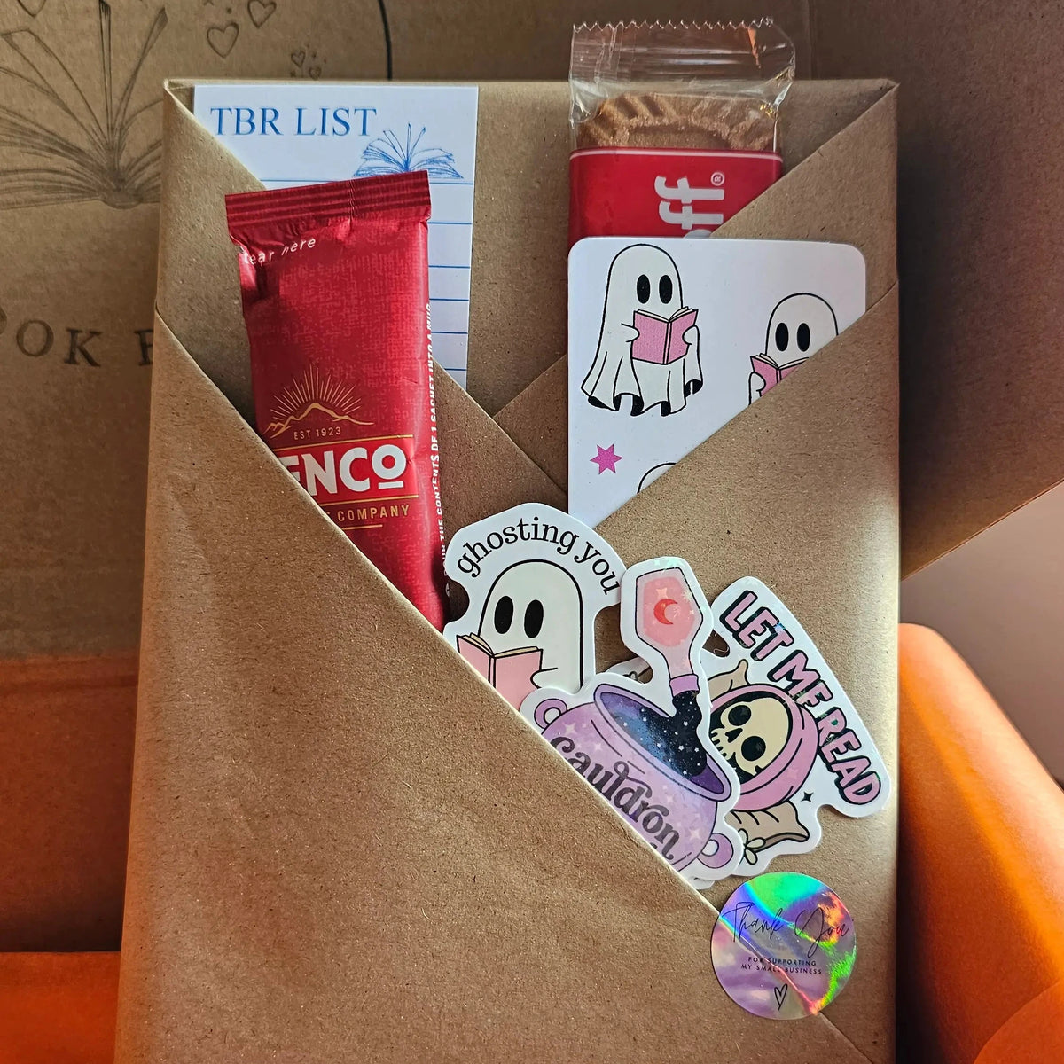 "Close-up of Spread Those Pages Halloween book box blind date with a book, with a pumpkin spiced latte sachet, biscoff biscuit, a ghost bookmark, a TBR (To Be Read) card and halloween themed bookish stickers
