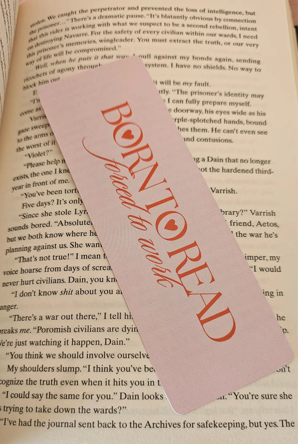  "Pink romance bookmark reading 'Born to Read, Forced to Work' lying across open romantasy book pages"