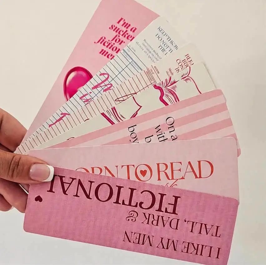 "Hand holding a fanned set of 7 pink romance bookmarks with bookish quotes and illustrations"