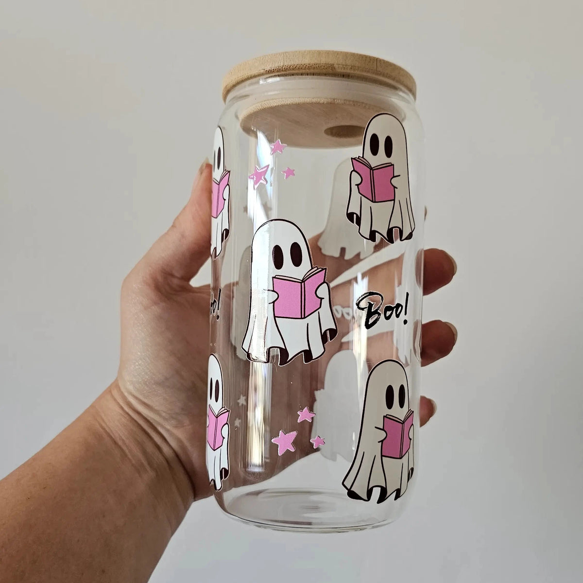 A hand holding a clear glass tumbler with a light wood lid. The glass features a repeating pattern of cute ghosts reading books and the word 'Boo!' printed in black script.